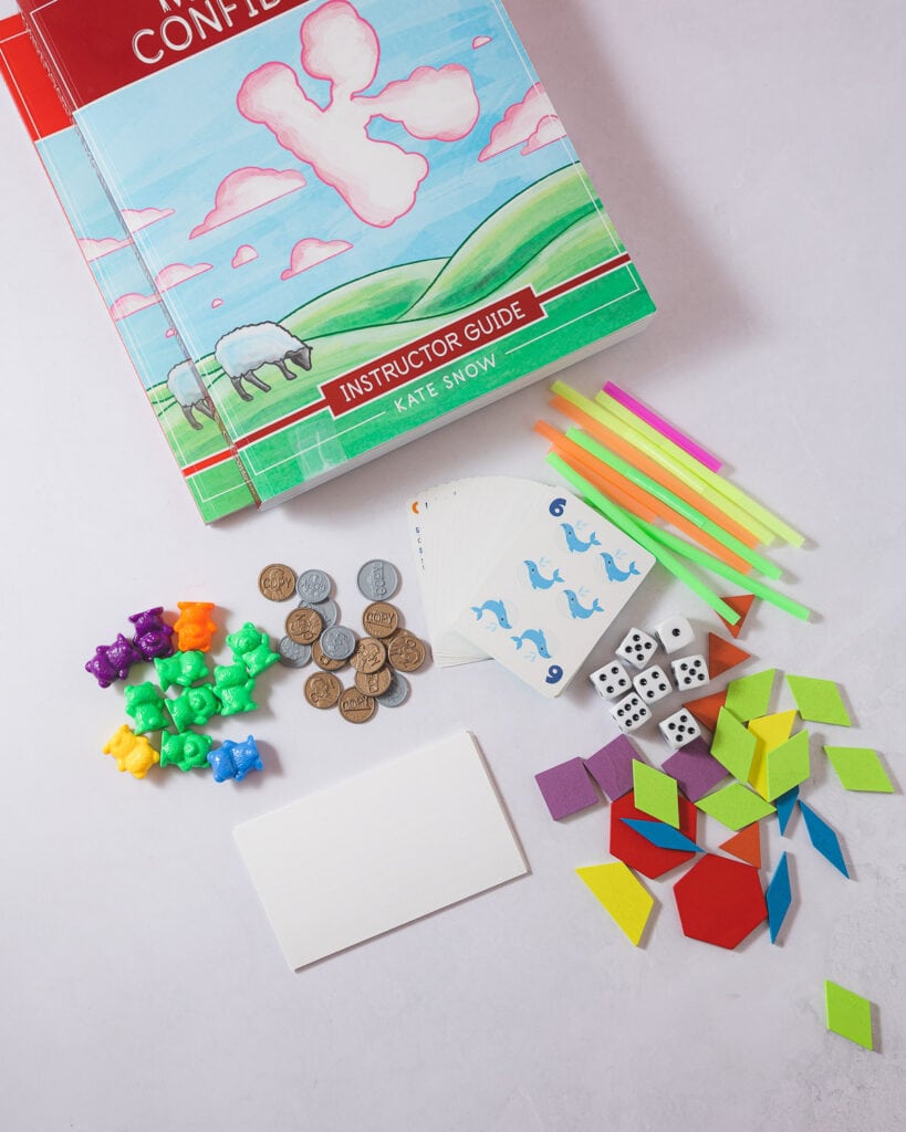 A selection of math manipulatives for kindergarten: counting bears, plastic play money, a deck of cards, index cards, dice, straws, and colorful pattern blocks.