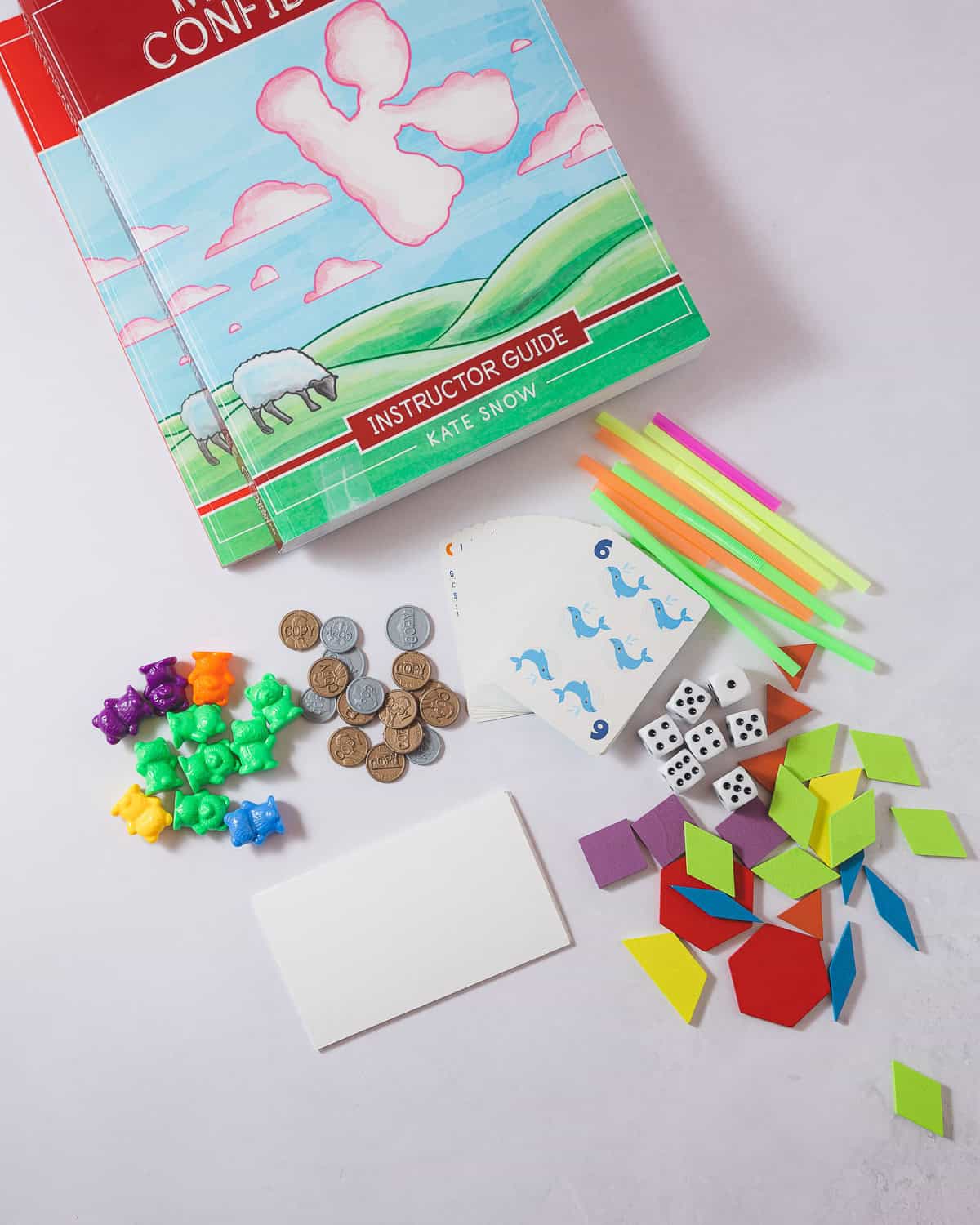 A selection of math manipulatives for kindergarten: counting bears, plastic play money, a deck of cards, index cards, dice, straws, and colorful pattern blocks.