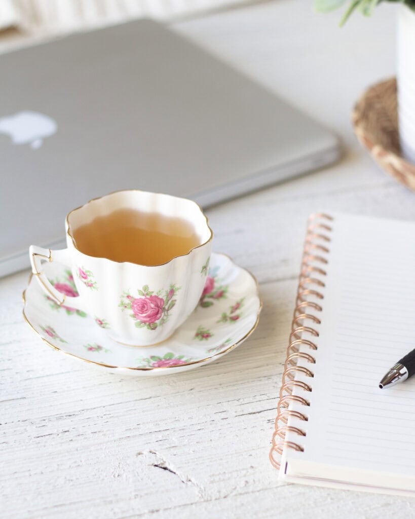 A floral teacup with tea in it next to a notebook with a pen.